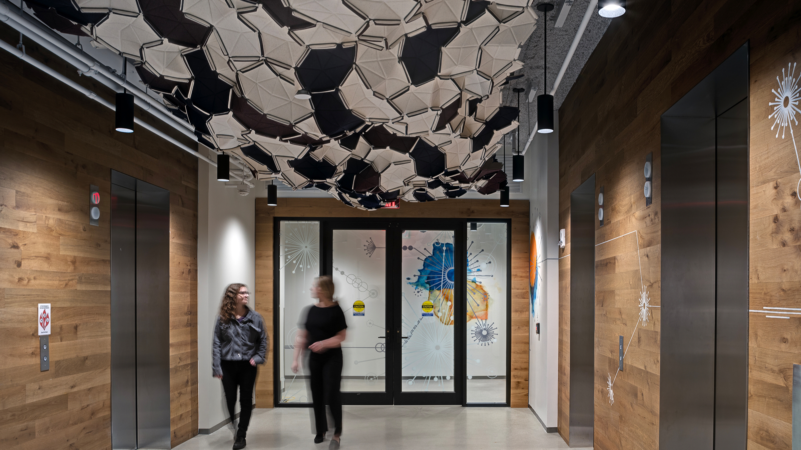 Two women in a motion time-lapse walking down a hallway with steel wire art on the ceiling